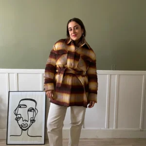 Une femme habillée d'un manteau à carreaux aux tons chauds (rouge, jaune, marron), porté avec un pantalon blanc et des baskets beiges. Elle est dans une pièce lumineuse avec un cadre décoratif moderne au mur.