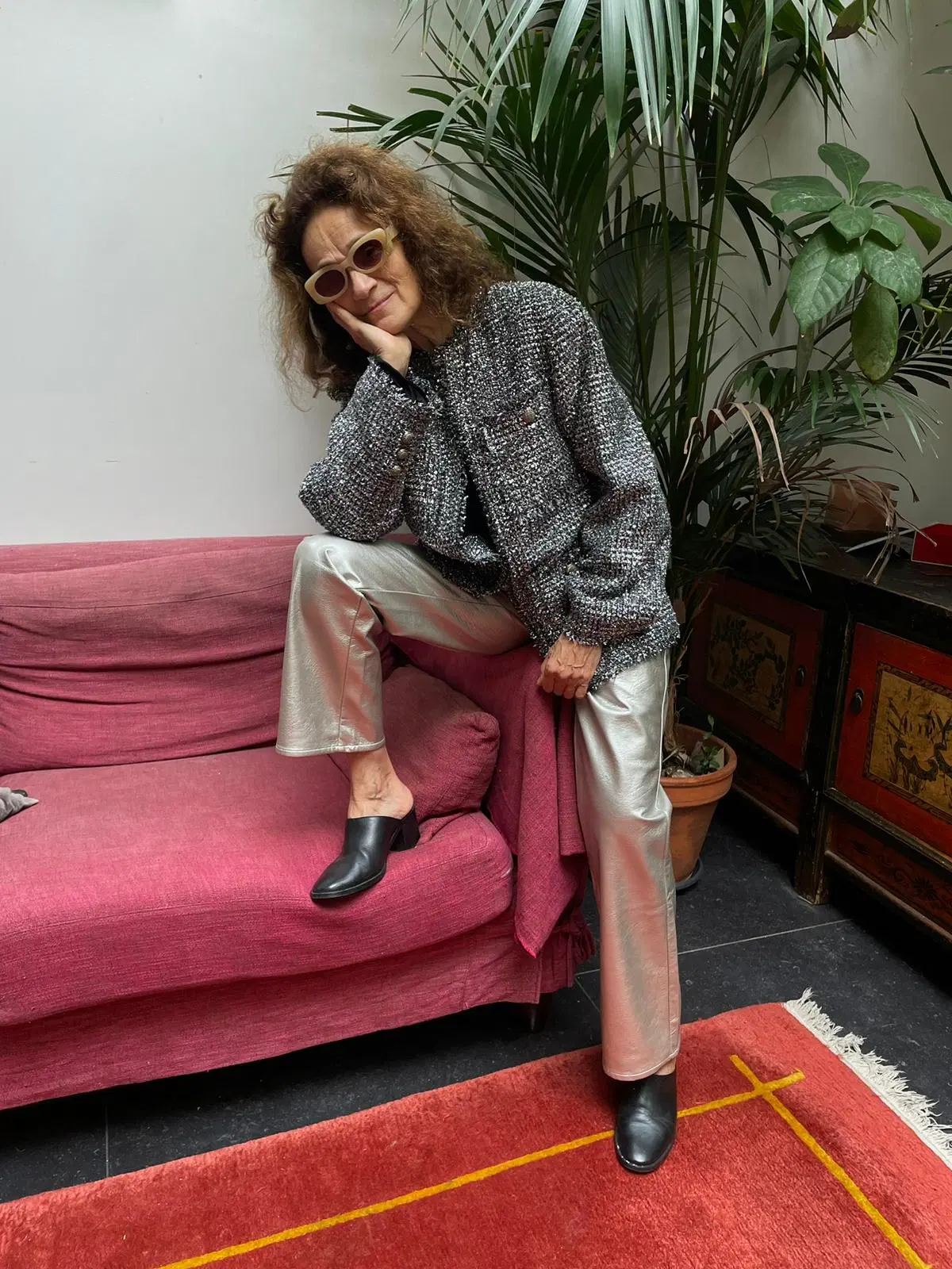 Femme posant dans une tenue élégante, portant une veste texturée noire et blanche avec des lunettes de soleil et un pantalon argenté en cuir vegan, assise sur un canapé rose devant des plantes vertes