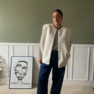 Une femme portant une veste sans manche molletonnée Urban Revivo, une chemise à carreaux, un t-shirt à maches longues rayés et un jean. Elle est debout dans une pièce lumineuse, avec un tableau minimaliste en arrière-plan.