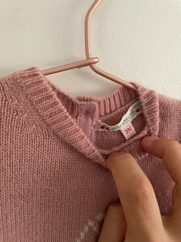 Combinaison pour bébé en tricot rose poudré, ornée d’un motif cerises blanches au centre. Marque bonpoint, 100%cachemire. suspendue à un cintre en cuivre