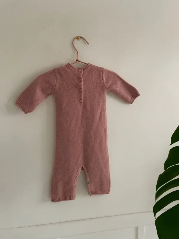 Combinaison pour bébé en tricot rose poudré, ornée d’un motif cerises blanches au centre. Marque bonpoint, 100%cachemire. suspendue à un cintre en cuivre